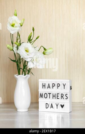 Scatola di luce con scritta Happy Mother's Day, fiori bianchi Eustoma o Lisianthus in vaso su sfondo di legno con spazio di copia. Foto Stock