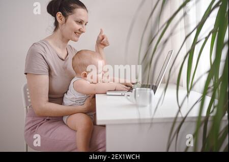 Giovane madre lavora studiando da casa con il computer portatile durante la quarantena, piccolo bambino carino sul giro. Ufficio domestico, genitorialità. Lavoro remoto, di Foto Stock