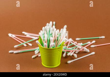 Secchio piccolo e bastoncini di cotone su fondo marrone. Un secchio verde pieno di bastoni sanitari. I bastoncini bianchi, verdi e rosa sono sparsi sullo sfondo Foto Stock