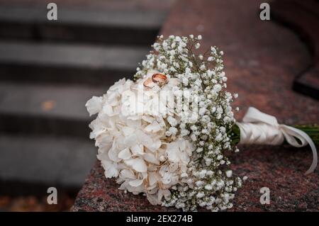 Arrangiamento floreale con fiori bianchi, bouquet di nozze con un nastro e due anelli d'oro, sdraiati su una pietra. Decorazioni per matrimoni, opere d'arte, fioraio Foto Stock