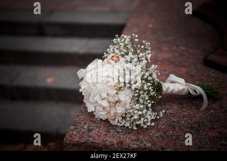 Arrangiamento floreale con fiori bianchi, bouquet di nozze con un nastro e due anelli d'oro, sdraiati su una pietra. Decorazioni per matrimoni, opere d'arte, fioraio Foto Stock