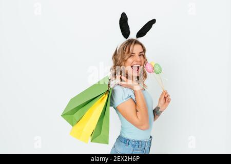 Festeggiando lo shopping di Pasqua, giovane donna che tiene le uova decorative colorate e i sacchetti di carta mentre si levano in piedi contro lo sfondo bianco sorridente e verricello. Foto Stock