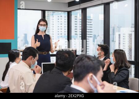 La donna esecutiva caucasica che mostra i pollici in su con propone la politica dell'azienda sull'uso della maschera di faccia in azienda durante la riunione. Nuovo ufficio normale. Mentre panne Foto Stock