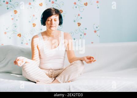 Una donna di mezza età si siede su un divano in posizione lotus con cuffie che ascoltano musica o lezioni di pratica spirituale su un computer portatile. Pratica di yoga Foto Stock