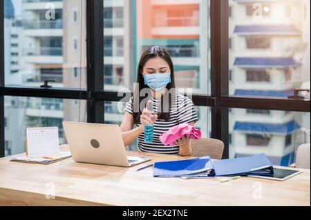 Donna dipendente indossare maschera per il viso preparare spray e panno imbevuto di alcol per la pulizia su scrivania in legno in ufficio. Dopo la riapertura e il blocco dell'azienda Foto Stock