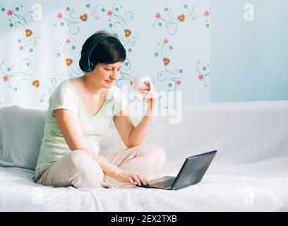 Felice donna di mezza età si siede sul divano con le cuffie, ascolta la musica, usa un computer portatile e bevande caffè o tè. Apprendimento a distanza lavoro intrattenimento Foto Stock