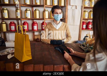 Giovane uomo in maschera medica che accetta il pagamento da parte del cliente Foto Stock