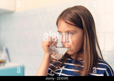 Ritratto di bambina che beve in ambiente domestico. Una bambina beve acqua da una tazza di vetro. Sete. Foto Stock