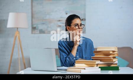 giovane donna pusiva che tiene la matita e guarda lontano vicino al computer portatile e libri sulla scrivania Foto Stock