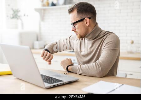 Un giovane imprenditore o insegnante affollato in occhiali seduti a casa e lavorando su un computer portatile, controllando il tempo da un orologio sul polso, il tempo per iniziare una conferenza online in streaming o una lezione virtuale Foto Stock