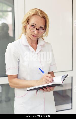 Donna con bicchieri e un cappotto bianco scrive in un notebook Foto Stock