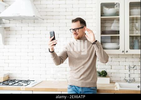 Bell'uomo hipster giovane bearded in occhiali in piedi in cucina e salutare un amico con high-five, guardando lo schermo, videochiamando via telefono cellulare, parlando, sventolando e dicendo ciao Foto Stock