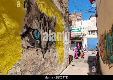 Marocco, Essaouira, scena di strada, vicolo con la faccia di gatto dipinto e donna che cammina oltre sulla strada Foto Stock