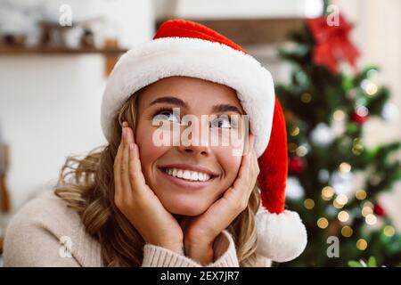 Primo piano di una giovane donna sorridente che indossa babbo natale cappello che guarda in su con christn=mas albero su uno sfondo all'interno Foto Stock