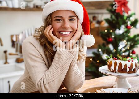 Donna felice nel cappello di babbo natale sorridente mentre si posa con Torta di Natale in una cucina accogliente Foto Stock