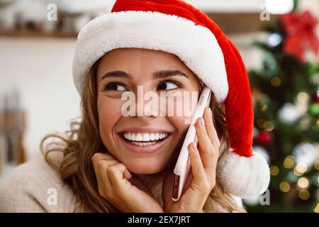 Felice bella donna nel cappello di babbo natale sorridente mentre parla sul telefono cellulare in una cucina accogliente Foto Stock