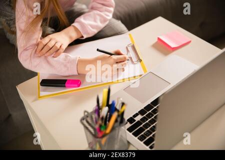 Chiudi le mani della ragazza che scrive durante la videochiamata di gruppo, utilizza la videoconferenza con l'insegnante, ascoltando il corso online. Utilizzo di cancelleria, notebook. Concetto di utilizzo facile, formazione, online, infanzia. Foto Stock