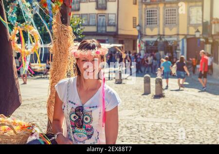 Giovane donna viaggiatore con corona di fiori guardando la macchina fotografica, sorridendo e posando vicino a stand souvenir Foto Stock