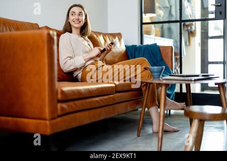 Bella donna con smartphone seduto sul divano marrone a casa mentre sorride e guarda la finestra. Stile di vita e concetto di lavoro da casa. Foto Stock