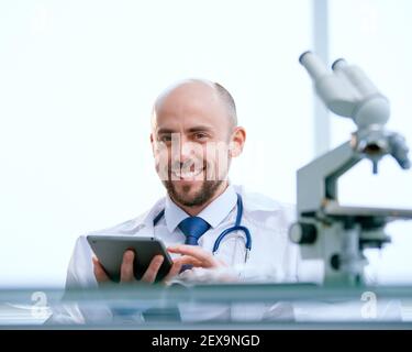 sorridente ricercatore con un tablet digitale seduto di fronte un microscopio Foto Stock