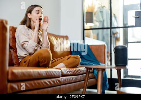 Felice bella donna che parla con la colonna nera dell'assistente audio sul tavolino da caffè mentre si siede sul divano di pelle marrone in un elegante soggiorno. Concetto di casa intelligente. Foto Stock