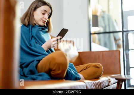 Bella donna con smartphone coperto da una plaid blu seduta sul divano marrone a casa. Stile di vita e concetto di intimità domestica. Foto Stock