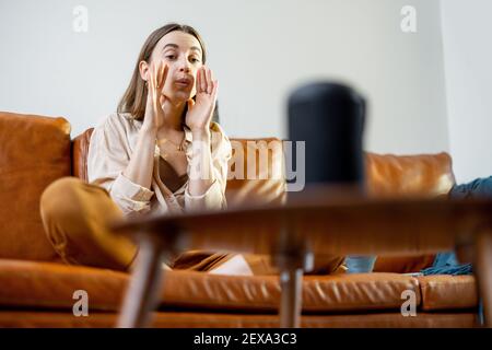 Felice bella donna che parla con la colonna nera dell'assistente audio sul tavolino da caffè mentre si siede sul divano di pelle marrone in un elegante soggiorno. Concetto di casa intelligente. Foto Stock