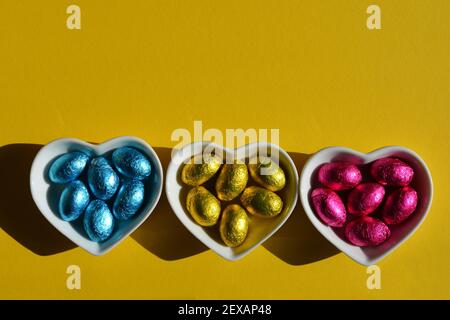 Uova di Pasqua avvolte in fogli blu, oro e rosa in piatti bianchi a forma di cuore in ceramica isolati su sfondo giallo Foto Stock