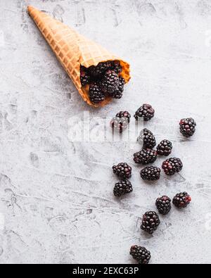 Still life flatlay di bacche nere fresche che fuoriescono da un cono di gelato alla cialda su una superficie blu chiaro macchiata. Foto Stock