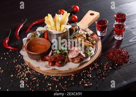 Assortimento di spuntini da salsicce, ali di pollo, patatine fritte, crostini, salsa su un piatto di legno su un tavolo di legno scuro. Alcol rosso in pile aroun Foto Stock