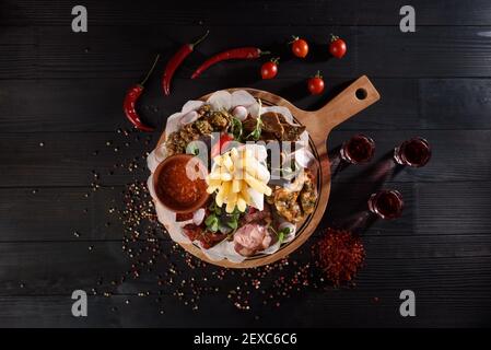 Assortimento di spuntini da salsicce, ali di pollo, patatine fritte, crostini, salsa su un piatto di legno su un tavolo di legno scuro. Alcol rosso in pile aroun Foto Stock