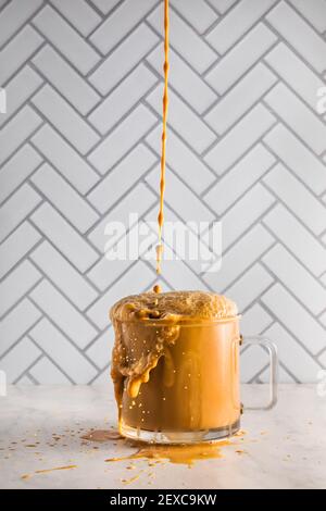 Il caffè ghiacciato cade e si spruzzi in una tazza di caffè in vetro traboccante che si trova di fronte a una superficie di piastrelle e sopra il marmo. Foto Stock