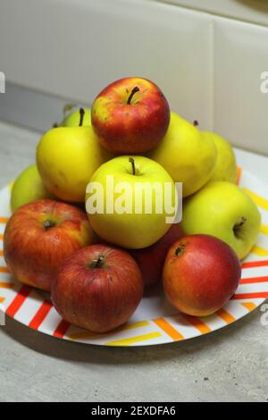 Un mucchio di mele in un piatto sul tavolo. Le mele mature rosse e verdi succose giacciono in un vetrino su un piatto bianco sullo sfondo di una parete grigia. Foto stock frutta vista dall'alto Foto Stock