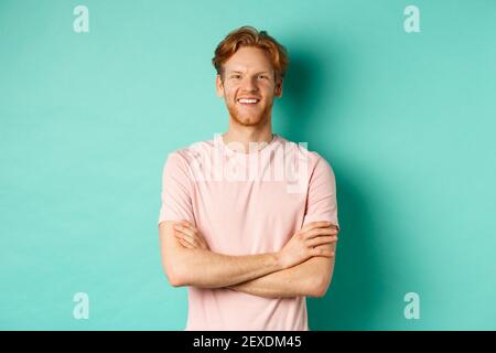 Ritratto di giovane dall'aspetto amichevole con capelli rossi e barba sorridente e appagata, tenendo le mani incrociate sul petto, in piedi sopra il turchese Foto Stock