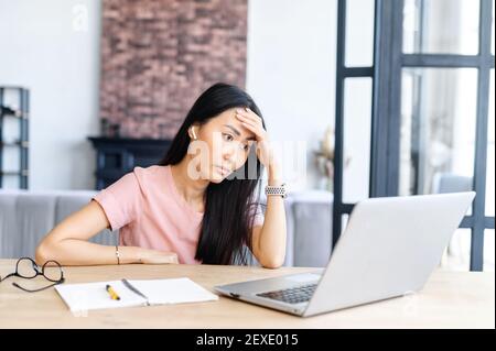 Stanca studentessa asiatica si sente annoiata e stanca, una bella donna brunetta guarda lo schermo del portatile senza entusiasmo, appoggiata testa sulla mano Foto Stock