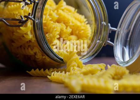 La pasta di Fussili cade da un grande vaso di vetro ermetico. Pasta secca cruda in contenitore di vetro a rifiuti zero. Composizione di cibo sudicio su sfondo scuro con Foto Stock