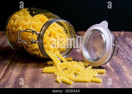 La pasta di Fussili cade da un grande vaso di vetro ermetico. Pasta secca cruda in contenitore di vetro a rifiuti zero. Composizione di cibo sudicio su sfondo scuro con Foto Stock