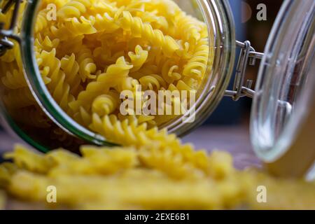 La pasta di Fussili cade da un grande vaso di vetro ermetico. Pasta secca cruda in contenitore di vetro a rifiuti zero. Composizione di cibo sudicio su sfondo scuro con Foto Stock