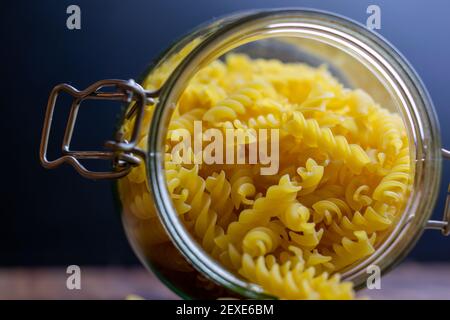 La pasta di Fussili cade da un grande vaso di vetro ermetico. Pasta secca cruda in contenitore di vetro a rifiuti zero. Composizione di cibo sudicio su sfondo scuro con Foto Stock