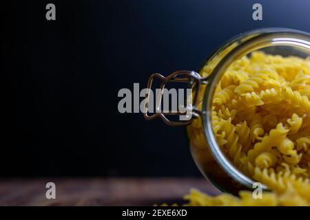 La pasta di Fussili cade da un grande vaso di vetro ermetico. Pasta secca cruda in contenitore di vetro a rifiuti zero. Composizione di cibo sudicio su sfondo scuro con Foto Stock