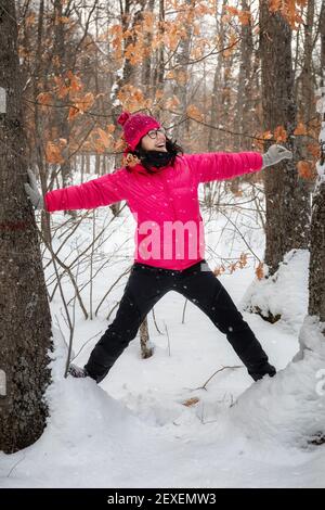 Donna felice nella foresta innevata, sorride e alza le braccia per la gioia della nevicata. Foto Stock
