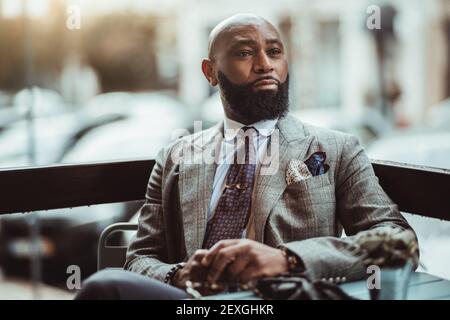 Ritratto di un imprenditore nero senza peli del dapper maturo con una bella barba, in un elegante costume grigio, seduto in un caffè all'aperto su una piovosa Foto Stock