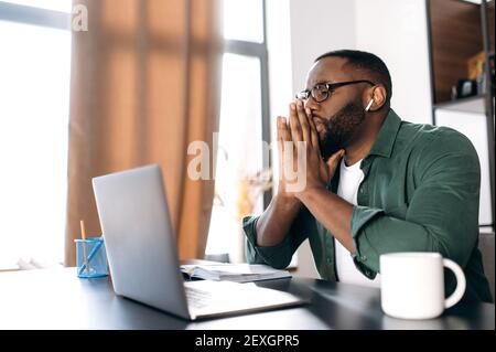 Serio uomo d'affari afro-americano focalizzato o freelancer che lavora su un progetto importante mentre sedendosi alla sua scrivania si concentra guarda in un portatile, pensando al lavoro Foto Stock