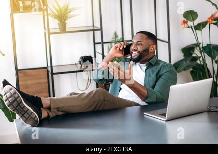 Felice Africano Americano giovane uomo, freelancer o ceo moderno, si sta rilassando sul posto di lavoro con gambe gettate sul tavolo, ha una piacevole conversazione telefonica con un amico o ragazza Foto Stock