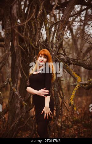 Donna ragazza dai capelli rossi in nero vestito foresta scura, ramo secco Foto Stock