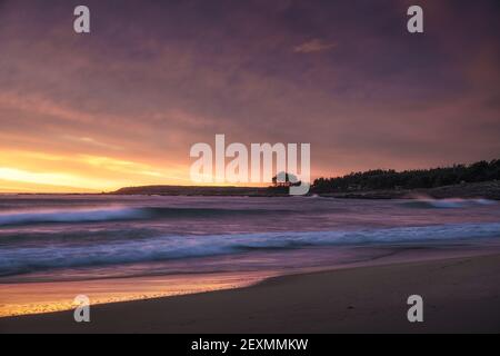 Una vista affascinante dell'oceano ondulato che colpisce la sabbia la spiaggia è splendente sotto il tramonto tra le nuvole Foto Stock