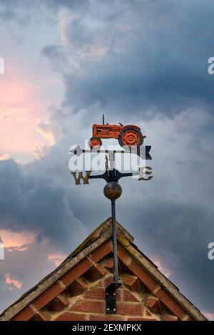 Gli agricoltori weathervane metallo rosso vecchio trattore sul tetto fienile mostra direzione nord est sud ovest direzione del vento con freccia verso l'interno davanti al cielo del tramonto Foto Stock