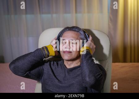 un uomo di 50 anni ascolta la musica con le cuffie a casa, seduto su una sedia. Delizia rilassante. Foto Stock