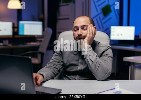 Un uomo d'affari assonnato che lavora su un computer portatile che fa straordinari sul posto di lavoro. Lavoratore dipendente che si addormenta a causa del lavoro di notte solo in ufficio per un importante progetto aziendale. Foto Stock