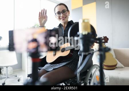 Donna in sedia a rotelle che tiene ukulele e che agita la mano alla macchina fotografica Foto Stock